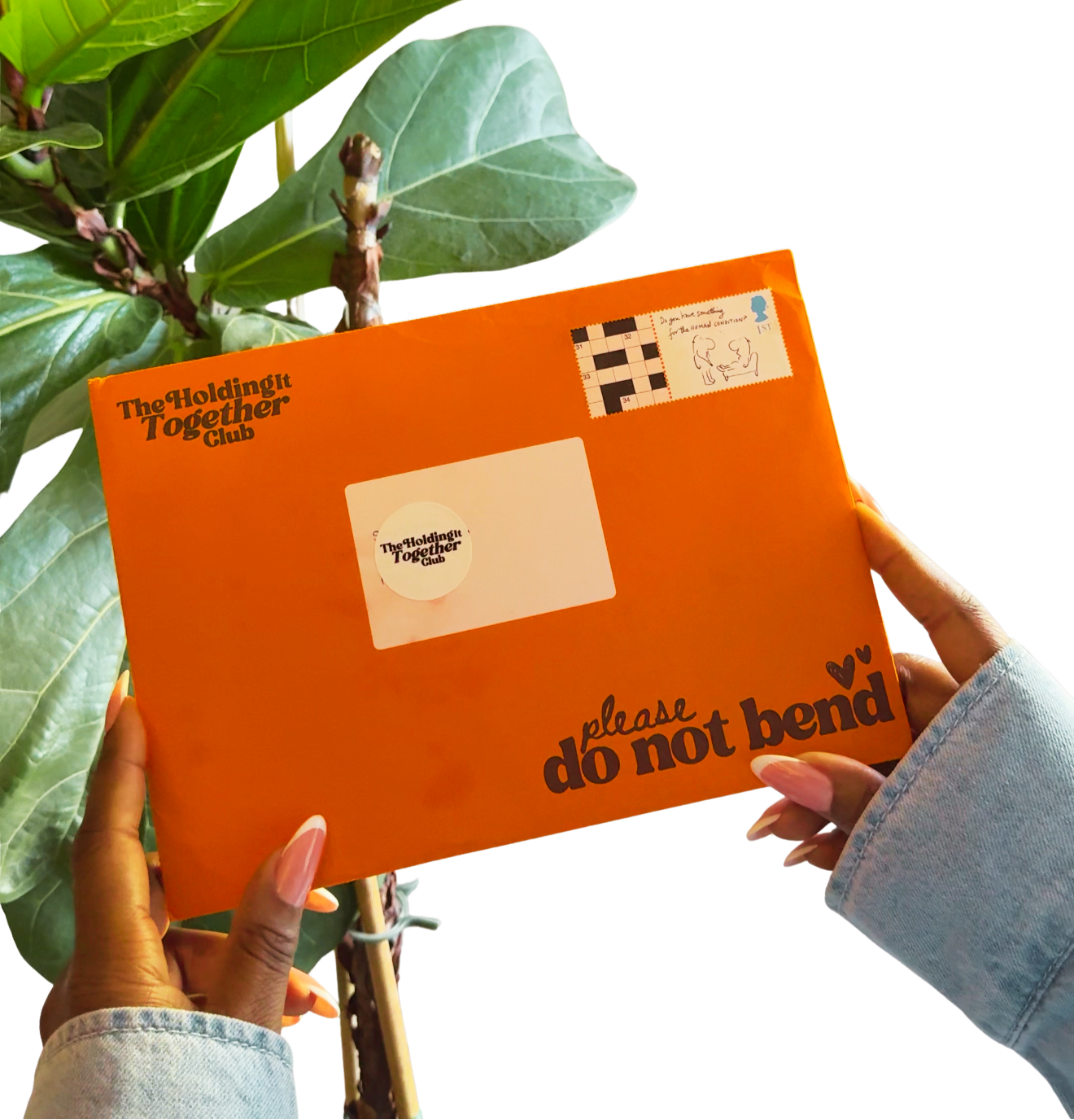 An orange envelope with 'The Holding Together Club' and 'please do not bend' printed on it, held by two hands against a white background with a green plant leaf in the corner.